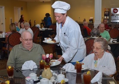 A chef in a white uniform serves food to an elderly man and woman seated at a dining table in a senior living facility's dining hall, with other residents dining in the background.
