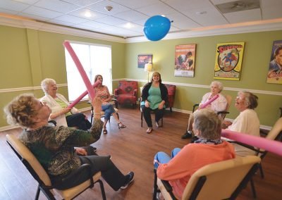 A group of elderly women sitting in a circle in a brightly lit room, engaging in a light exercise activity by batting a blue balloon with pink foam noodles, while two caregivers look on.