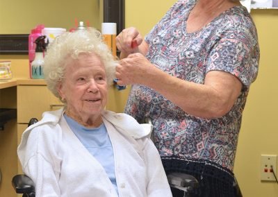 An elderly woman sitting in a chair and smiling as another woman styles her hair in a well-lit room, with various hair styling products on a counter in the background.