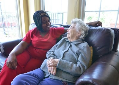 A caregiver in red scrubs sitting on a couch and warmly interacting with an elderly woman in a gray sweater, both smiling, in a room with large windows providing natural light.