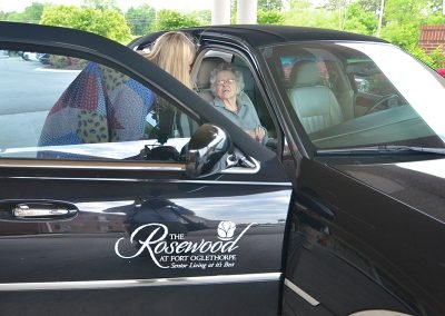 An elderly woman seated in the passenger seat of a black car with 'The Rosewood at Fort Oglethorpe' logo, while a caregiver standing outside the car leans in to assist her, in a parking area with greenery in the background.