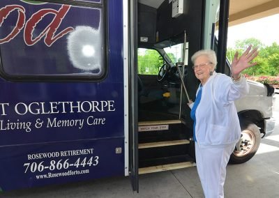 An elderly woman waving and smiling as she prepares to board a Rosewood at Fort Oglethorpe shuttle bus, with the bus door open and the interior steps visible.