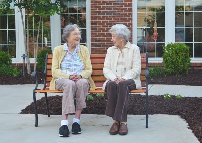 Two elderly women sitting on a bench outside a brick building, smiling and conversing with each other, surrounded by landscaped bushes and a bird feeder in the background.