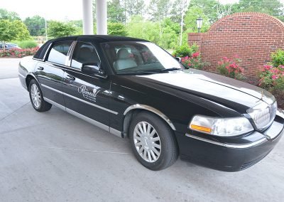 A black luxury car with 'The Rosewood at Fort Oglethorpe' logo on the door, parked under a covered driveway area, with a background of greenery and a brick wall.